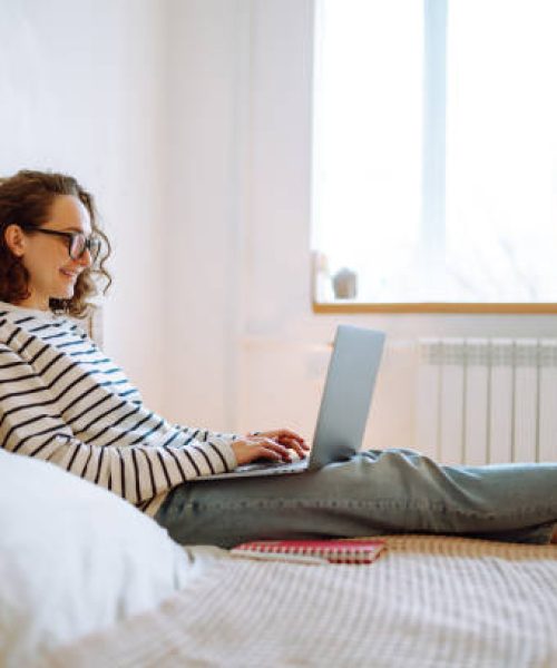 Young female freelancer works and makes notes while working at home in quarantine lockdown. Freelancer with wireless headphones calling on laptop, talk by webcam. Education,shopping and working online